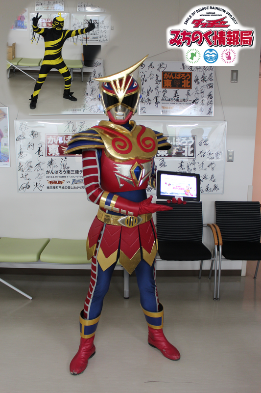 待合スペースで、赤と金の甲冑風の衣装にマスクを着けたたニューヒーロー『未知ノ国守ダッチャー』タブレットを手に正面を向いて立ち、左奥には黒と黄色の縞模様のケヤンキーもポーズをとって写っている写真