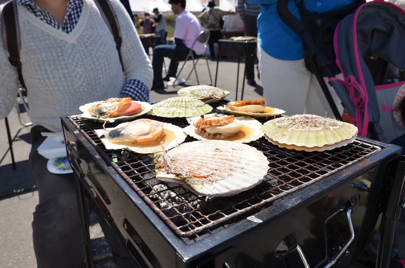 屋外の会場で殻付きのホタテが網の上に並べられ炭火で焼かれており、会場には多くの人が訪れ、ホタテまつりを楽しんでいる様子を写した写真