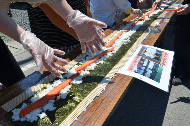 屋外の長いテーブルの上に海苔とご飯、しゃけが並べられ、ビニール手袋をつけた参加者たちが協力して長いのり巻きを作っている様子の写真