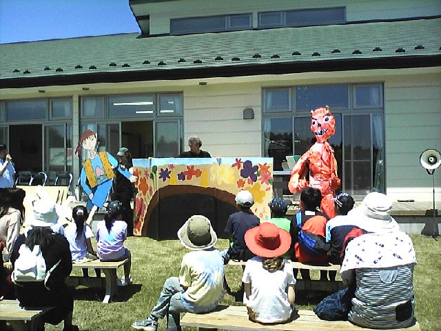 青空の下、コアラ館前の芝生広場で、親子連れの観客がベンチに座っており、前方では大きな人形を使った人形劇が行われ、楽しんで見ている様子の写真