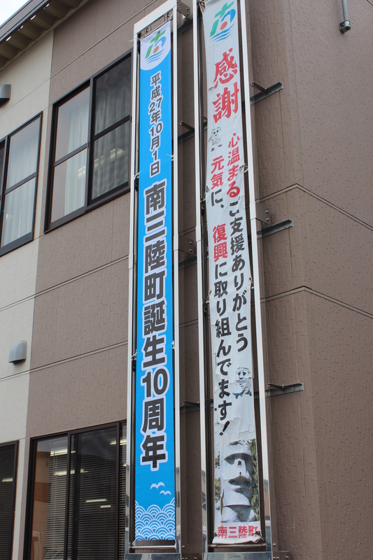 役場建物の壁面に左側には「南三陸町誕生10周年」と書かれた青い横断幕、右側には「感謝 心温まるご支援ありがとう 元気に復興に取り組んでます！」と書かれた白地の横断幕が並んで掲げられている様子を写した南三陸町誕生10周年の懸垂幕の写真