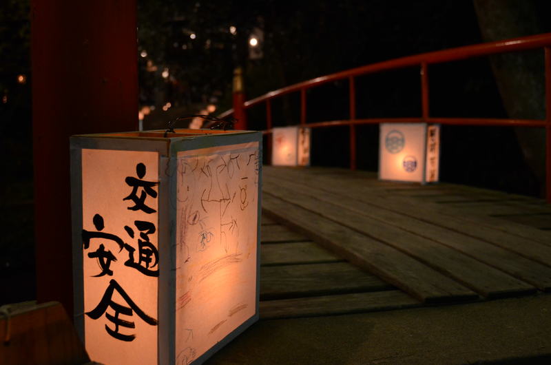 赤い欄干の木橋に沿って並べられた提灯が夜の闇を暖かく照らし、提灯には「交通安全」などの文字が書かれており、上山八幡宮の秋まつり（夜神楽）の静かで厳かな雰囲気を伝える夜の境内の写真