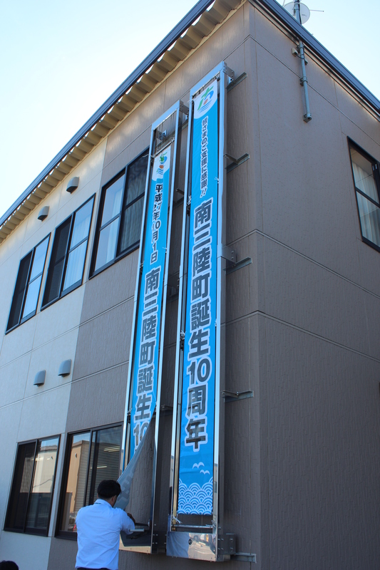 職員の男性が掲示作業をしており、役場建物の壁面に「南三陸町誕生10周年」と書かれた青い縦長の横断幕が二本並んで掲げられている様子の写真