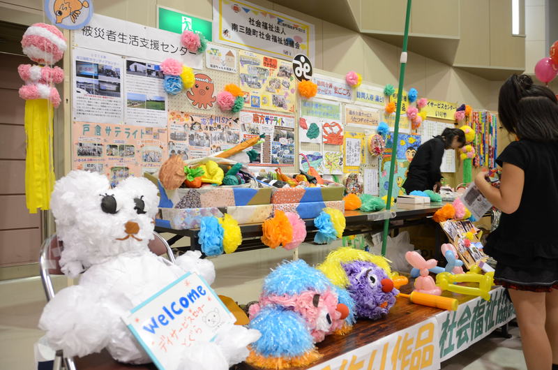 屋内の展示ブースに、白い手作りのテディベアが「Welcome」と書かれた紙を持って椅子に座り、テーブルの上にはカラフルな紙花やふわふわした人形、風船の動物などの手作り作品が並び、壁には「被災者生活支援センター」などの案内ポスターや写真が飾られ、右側で子どもが興味深そうに見ている地域福祉イベントの展示ブースの写真