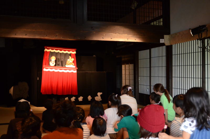 舞台で人形劇が行われており、多くの親子連れが座って人形劇を楽しんて見ている様子の写真