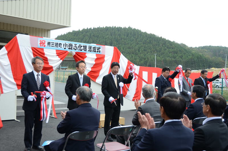 「祝 南三陸町 小森ふ化場 落成式」と書かれ紅白幕が張られ屋外の会場で、町長と関係者の方々がテープカットを行っており、多くの来場者が席に着き拍手をしている小森ふ化場落成式の様子の写真