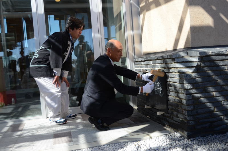 スーツ姿の男性が白い手袋をはめて木槌を使い、南三陸病院の定礎として建物の壁面に納める記念の石板を慎重にはめ込んでいる様子が写っており、その後ろで法被を着た男性がかがんで見守っている南三陸病院定礎式の様子の写真