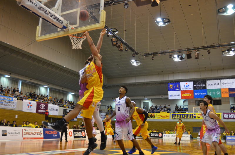 室内のバスケットボールコートで、黄色と紫のユニフォームを着た選手たちが、ゴール付近で競り合っている様子の写真