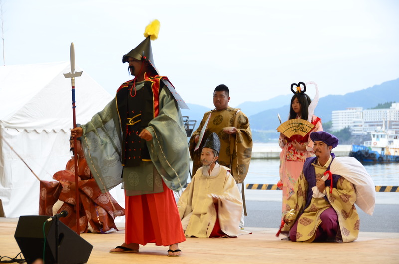 港を背景に、華やかな歴史衣装を着た6人の男女が屋外の舞台で本浜七福神舞を演じている様子を写した写真