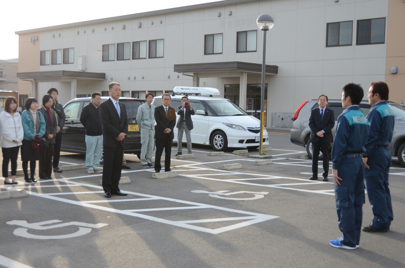 建物の前の駐車場で、作業服を着た二人の男性とスーツ姿の男性が向かい合って立ち、その後ろには複数の関係者の人々が整列している職員派遣出発式の写真