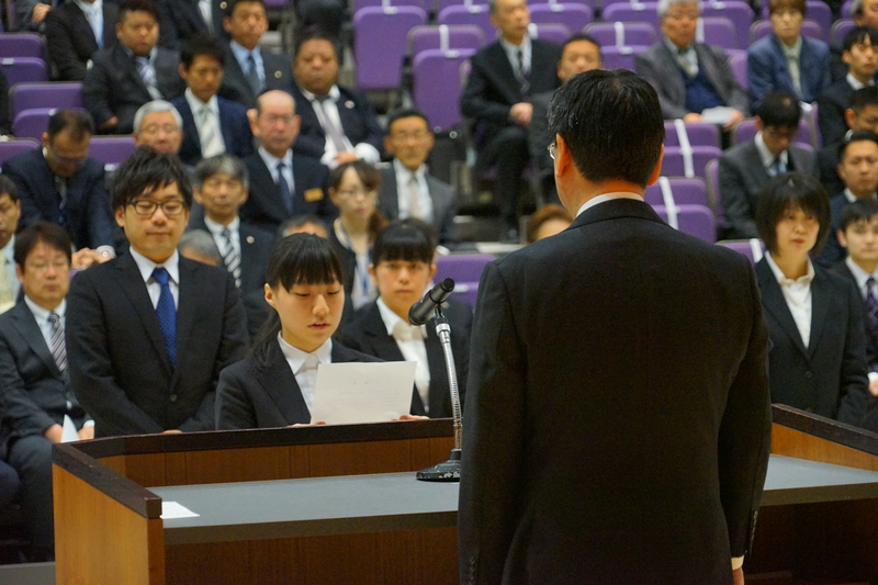 アリーナの座席に着席したスーツを着た大勢の人々が見守る中、若い女性が演台に立った男性に向かって文書を読み上げている厳かな場面の写真