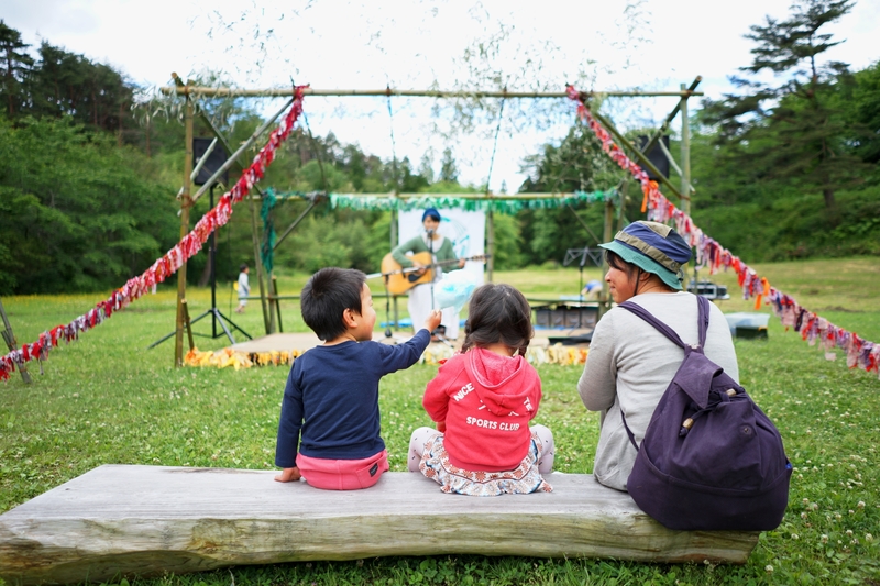 芝生の公園に設置された野外ステージでギターを弾く人を、ベンチに座った親子が楽しそうに見ている様子の写真