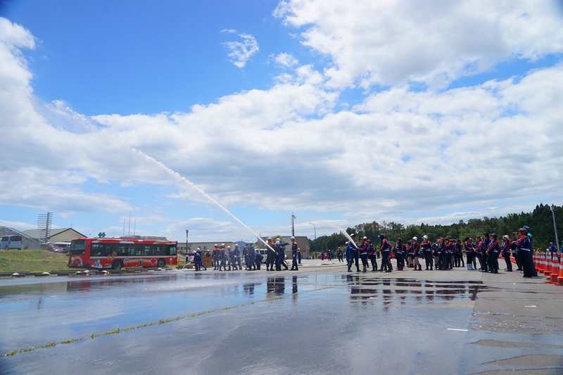 大勢の消防団員たちが青空の下で一列に並び、ホースから水を放って訓練している様子の写真