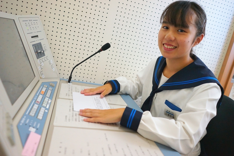 セーラー服を着た女子生徒が放送室のマイクの前で原稿に手を添え、笑顔でカメラを見ている写真
