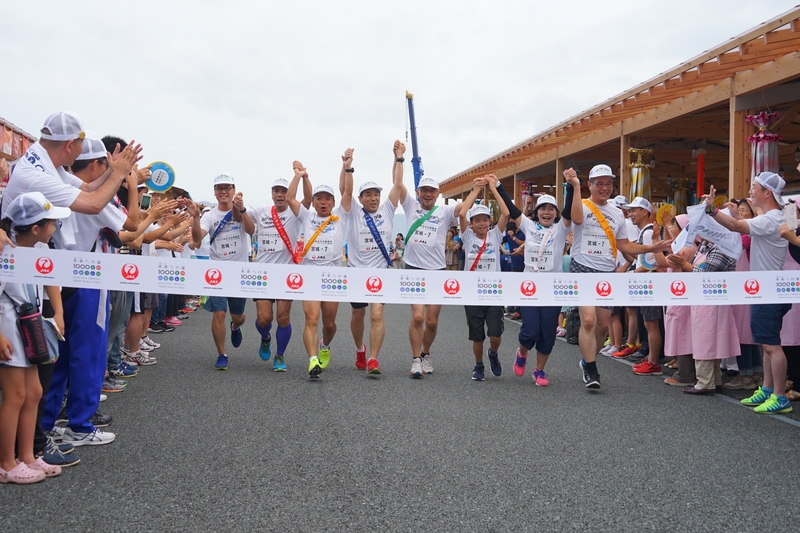 色とりどりのタスキをかけた8人ンのランナーたちが、繋いだ手を高く上げながら揃ってゴールテープを切ろうとしている瞬間を、沿道の人々が拍手で祝福している様子の写真