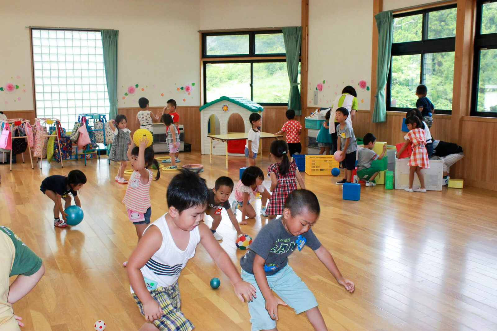 奥では保育士が数人の子どもたちと一緒に遊んでおり、明るい木の床と大きな窓に囲まれた室内で、多くの園児たちがボール遊びやおもちゃで自由に遊んでいる様子の写真