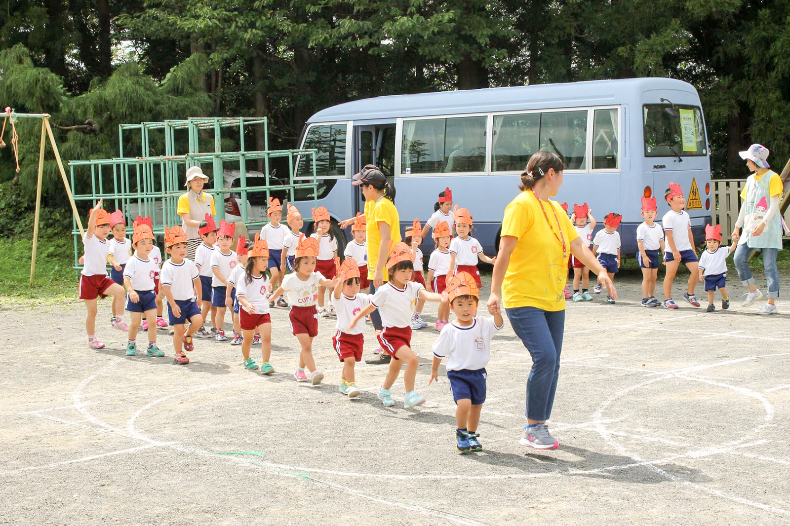 木々に囲まれた背景には園バスや遊具が設置された園庭で、数名の保育士がお揃いの黄色いTシャツを着て誘導するなか、赤やオレンジの動物のお面を頭にかぶり白い体操服と赤や青の短パンを着た園児たちが、女性の保育士さんを先頭に整列しながら行進している様子の写真