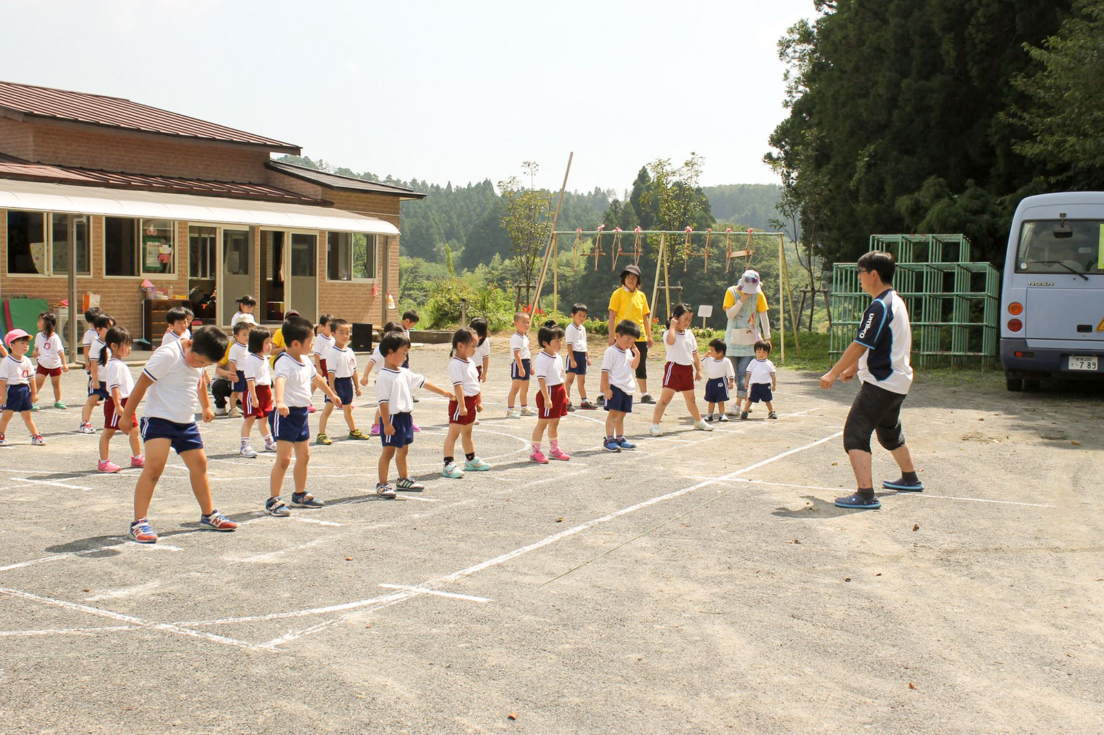 背景には遊具や送迎バスが見える、自然に囲まれた園舎の前の広い園庭で、白い体操服と赤や青の短パンを着た大勢の園児たちが間隔をあけて整列し、男性保育士の動きに合わせて体操のような運動をしている様子の写真
