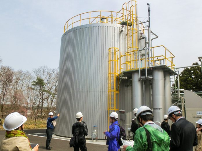 白い防護ヘルメットをかぶった複数の見学者が、屋外にある銀色の大きなタンク状のバイオガス施設の前で案内役の男性から説明を受けている様子の写真