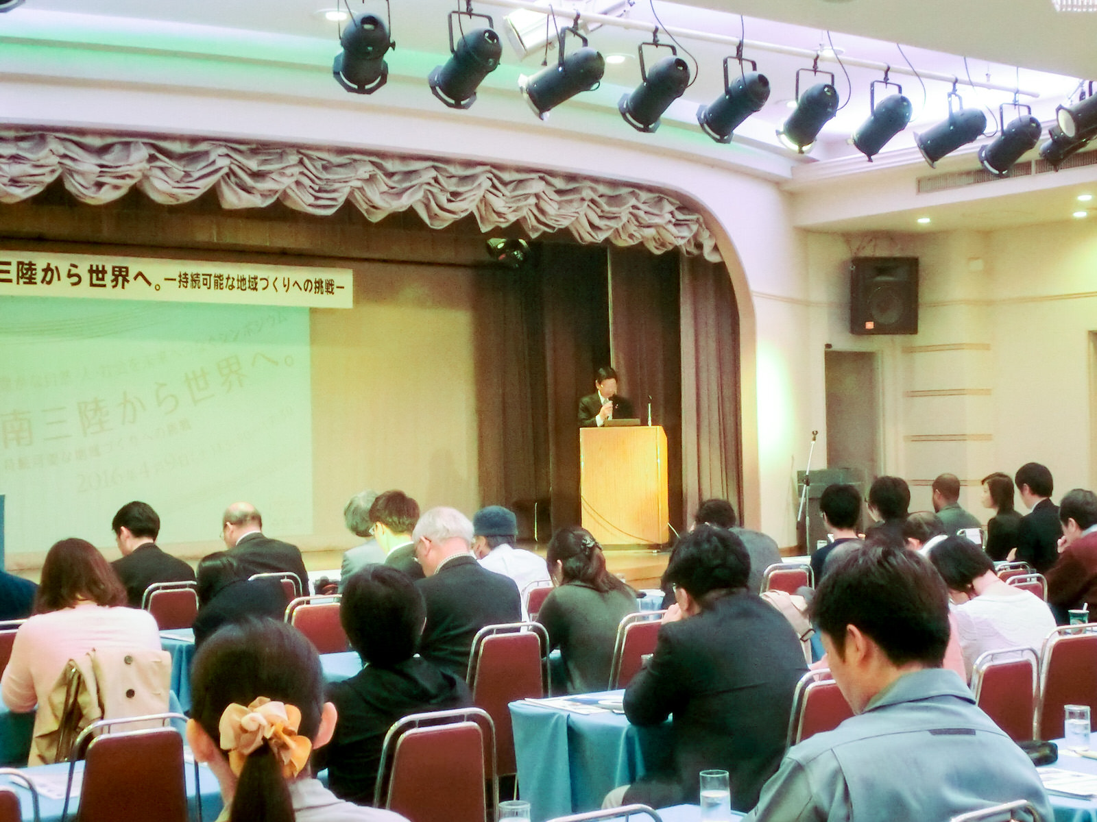 広い会場で、参加者が机に向かって座り、前方のステージ上の演台に立つ講演者の男性がマイクを使って講演している様子を後方から撮影した写真