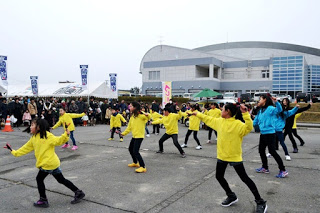 屋外の広いイベント会場で沢山の観客が見ているなか、黄色と青のジャケットを着た子どもたちが集団でよさこいを踊っている写真