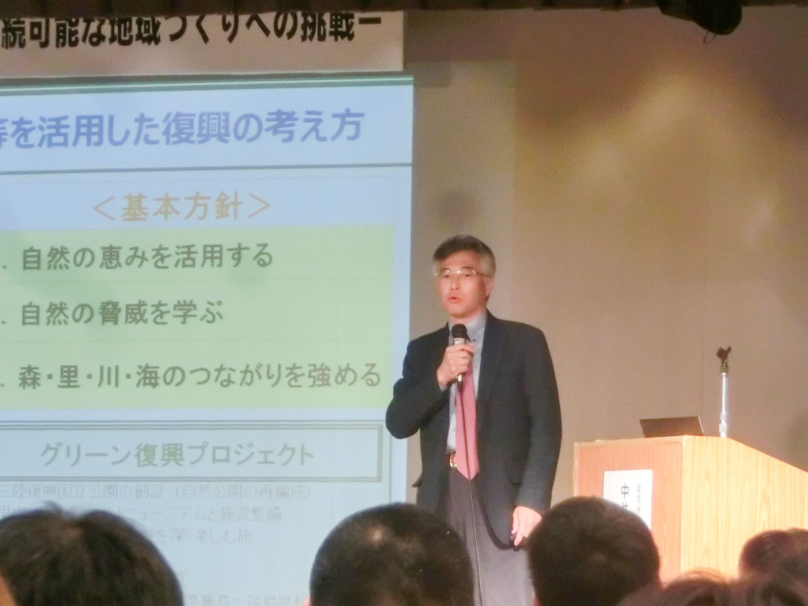 ステージの大きなスクリーンに資料が映し出され、その前に立った講演者の中井 徳太郎氏がマイクを持って話をしている写真