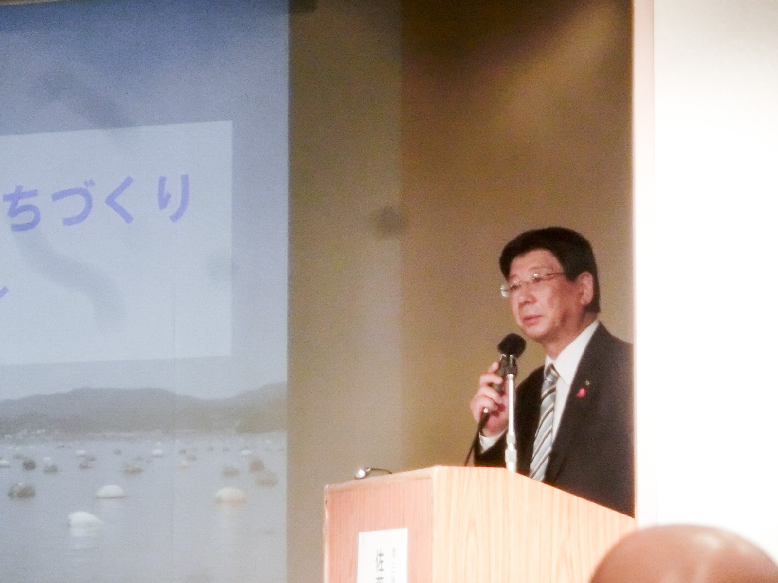 資料が映し出された大きなスクリーンの横にある演台に立ち、マイクを持って立っている佐藤町長の写真
