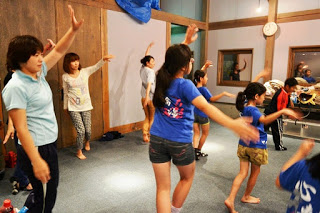 室内で数人の女性と子どもたちが、左手を上げ、右腕を横に真っすぐ伸ばしよさこいを踊っている写真