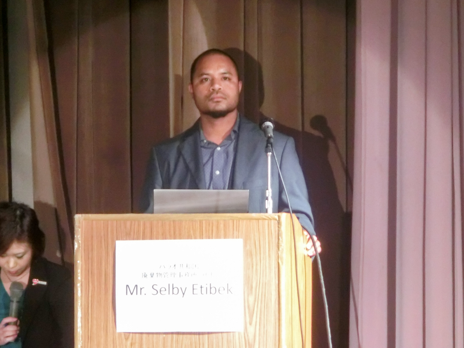 「Mr. Selby Etibek」と書かれた紙が貼られた演台にスーツを着た外国人の男性が立っており、その横にマイクを持った女性が座っている写真