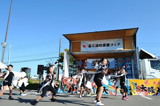 カラフルな大漁旗が飾られた屋外の舞台の前で、黒と白の衣装を着た子供たちが踊っている様子の写真