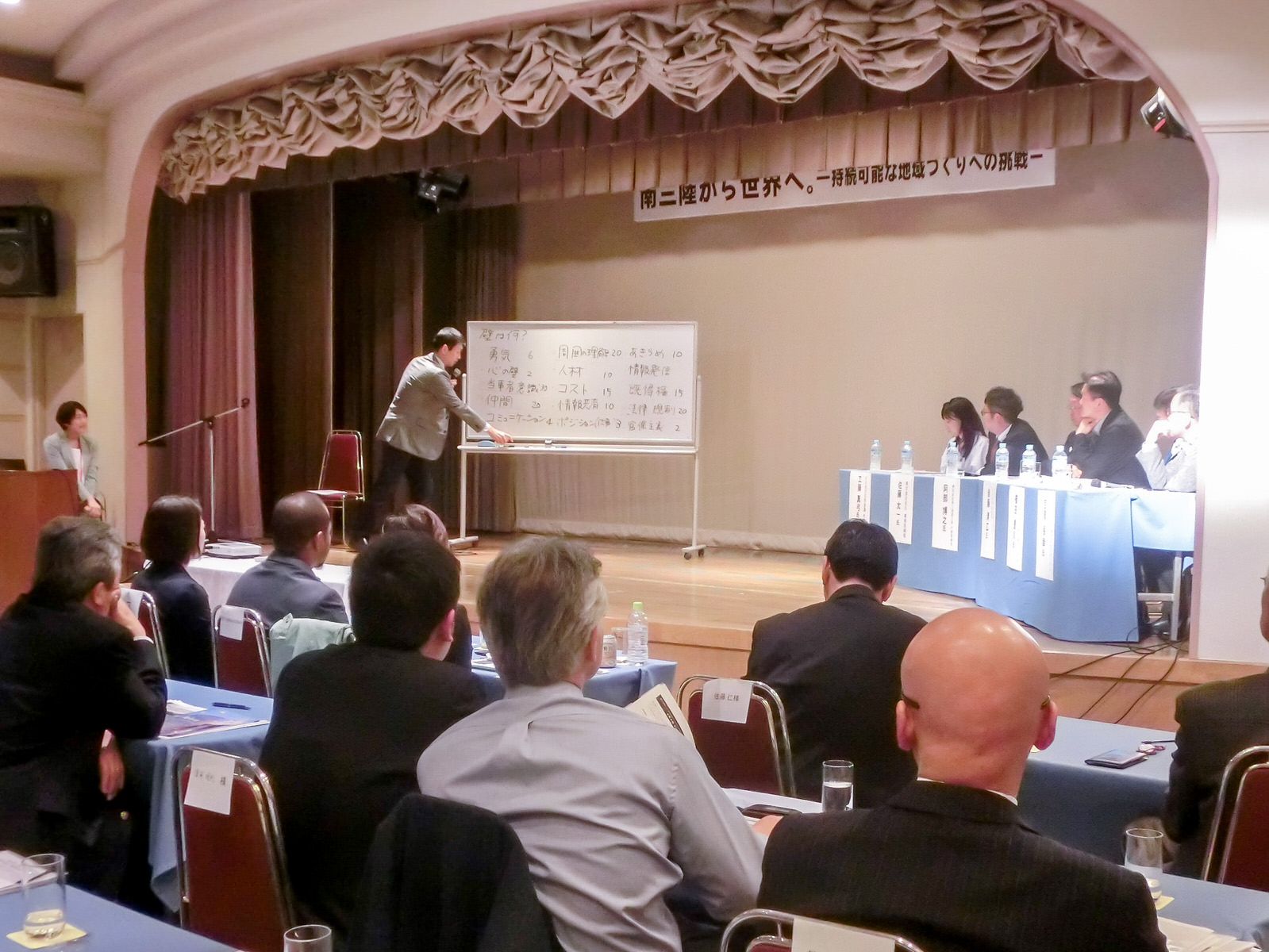 ステージ上でマイクを持った男性がホワイトボードの前に立ち、壇上に置かれたテーブルの席に座った男女数名と、客席に座った参加者たちがホワイトボードの方を見ている意見交換会の様子の写真
