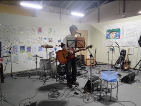 壁に多数の絵が飾られた室内で、アコースティックギターを弾きながらマイクの前に立ち歌っている男性の全身写真