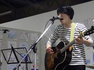 壁に複数の絵や作品が飾られた室内で、男性がアコースティックギターを弾きながらマイクの前に立って歌っている写真