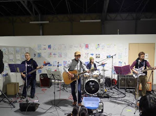 背景の白い壁にはさまざまな絵が貼られている室内で、中央のスタンドマイクの前で男性がアコースティックギターを演奏しながら歌を歌い、その左右に立った男性2人がエレキギターを弾き、後ろに座った男性がドラムを叩いている、バンド演奏中の様子の写真