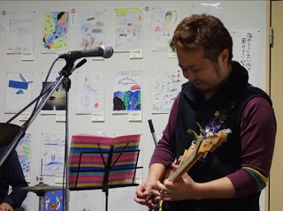 指でギターの弦を押さえながら笑顔で演奏しているいる男性の写真