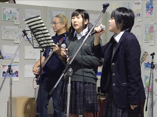 壁にさまざまな絵や作品が飾られた室内で、男性のギター演奏に合わせて、2人の女子高校生がマイクを持って歌っている写真