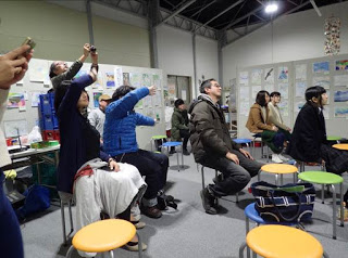 壁にさまざまな絵が飾られている部屋の中で、複数の観客がカラフルなスツールに座って演奏を楽しんでおり、後ろに立った2人がスマートフォンで撮影している様子の写真