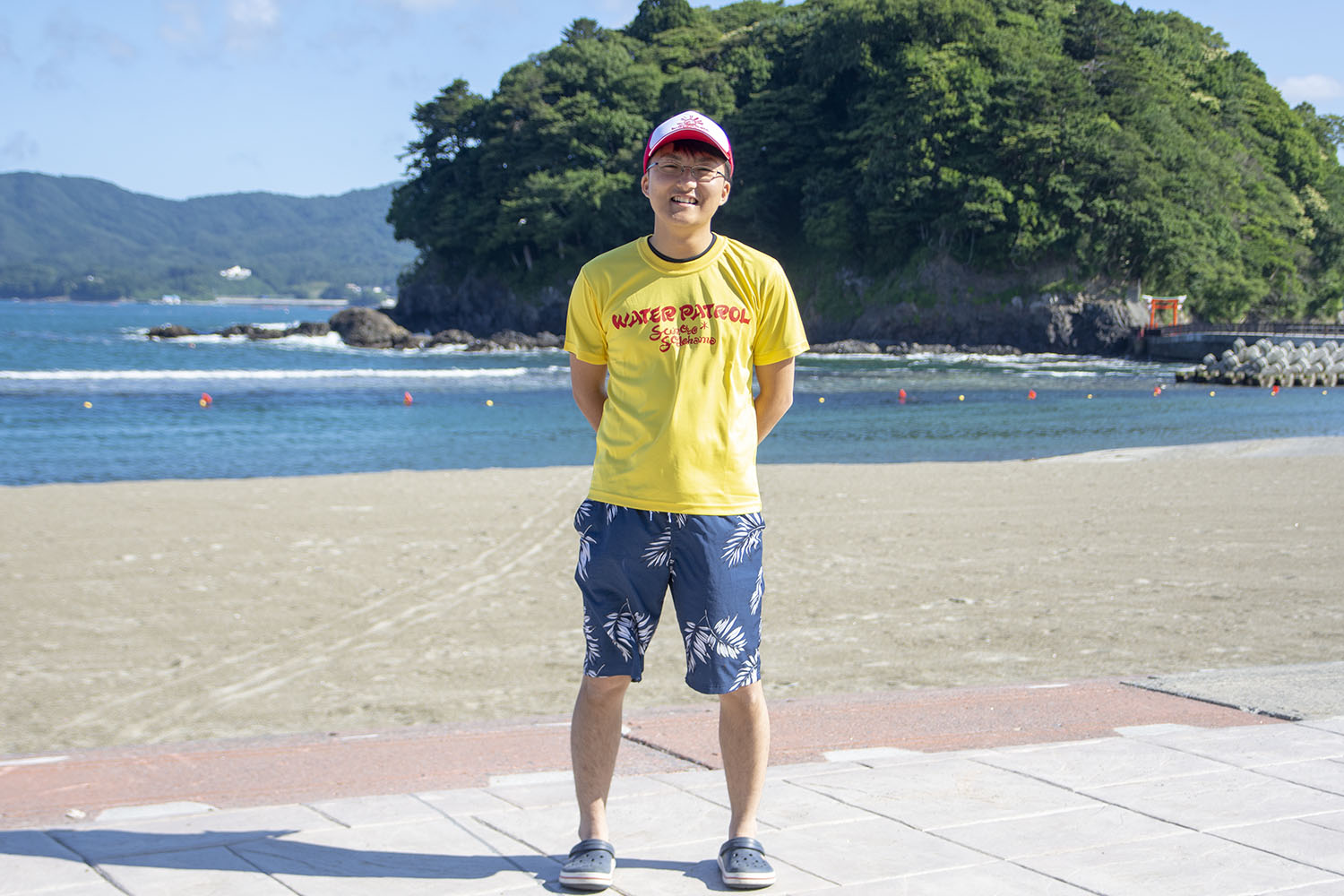 後方に荒島の見える海岸で黄色い「WATER PATROL」Tシャツと青い花柄の短パン、赤と白の帽子をかぶった笑顔の阿部悠斗さんの写真