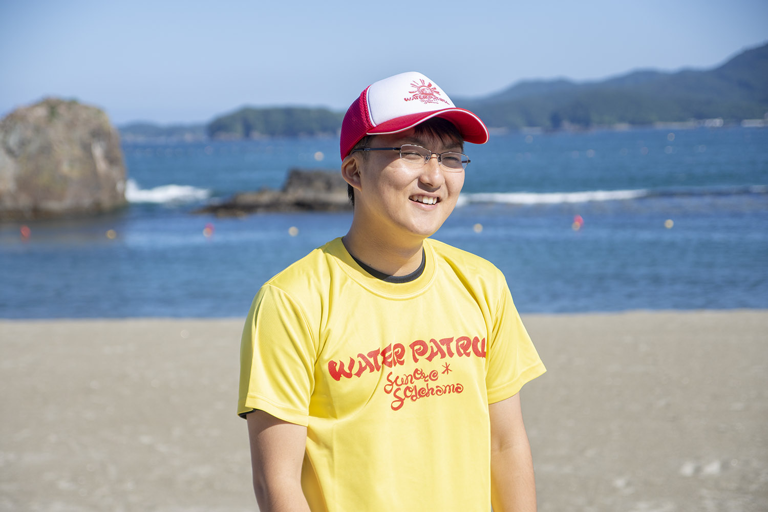 海辺で黄色い「WATER PATROL」と書かれたTシャツを着て、赤と白の帽子をかぶった阿部悠斗さんが笑顔で話をしている様子をアップで撮影した写真