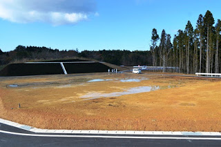 周囲を林に囲まれ、ところどころ水たまりのある整地済みの広い区画の奥に車と人が見える宅地の写真
