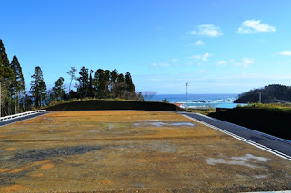 整地された広い区画の奥に黒い法面と海が見え、青空の下に水平線が広がっている造成地の写真