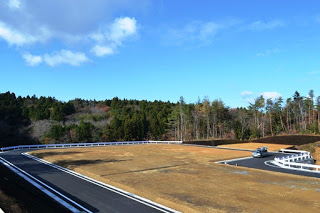 舗装された道路に囲まれた広い区画の奥に車が停まり、背景には木々が茂る山林がある造成地の写真