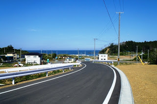 緩やかに曲がる舗装道路の先に海と住宅が見え、両側に電柱が立ち並ぶ坂道の写真