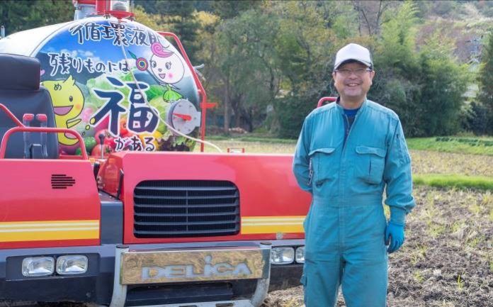 「残りのもには福がある」の文字と野菜がイラストされたキャタピラ車の横で水色の作業服を着た笑顔の西城勝志さんの写真