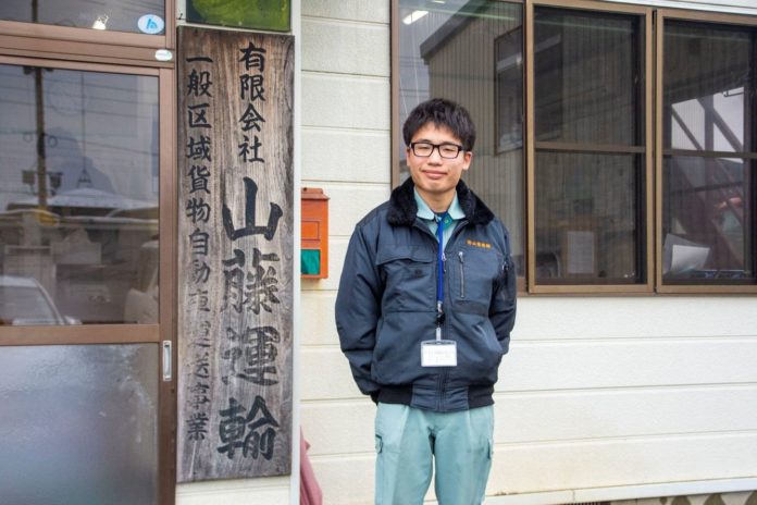 名札を首から下げて紺色のジャンパーを着た佐藤さんが、山藤運輸と書かれた木の看板の横で微笑んで立っている写真