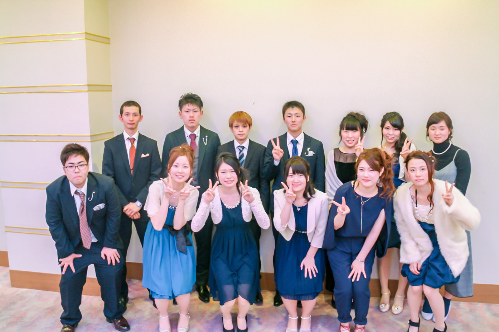 式典会場の一角で、スーツやドレス姿の男女13人が2列に並んでピースサインをしながら笑顔で記念撮影をしている写真