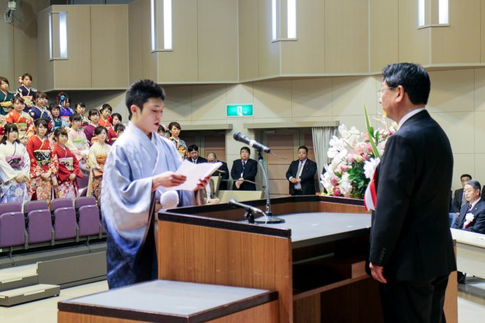 観客席では振袖姿の女性たちが整列している成人式の式典会場で、壇上に立つスーツ姿の佐藤町長が聞いている中、羽織袴を着た新成人代表の男性が原稿を読み上げている様子の写真