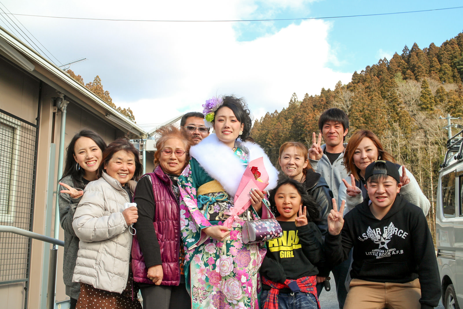 晴れた屋外で、振袖を着た若い女性が花束を持ち、家族や親族らしい9人が周囲を囲んで笑顔で集合写真を撮っている写真