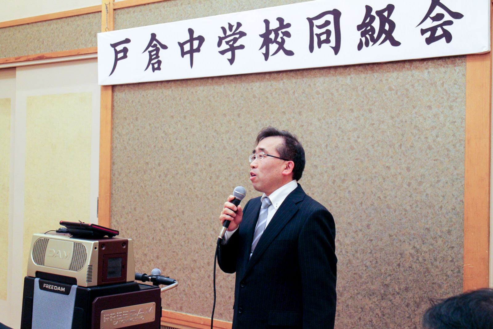 「戸倉中学校同級会」と書かれた横断幕の前で、スーツ姿の男性がマイクを持ってスピーチをしている写真