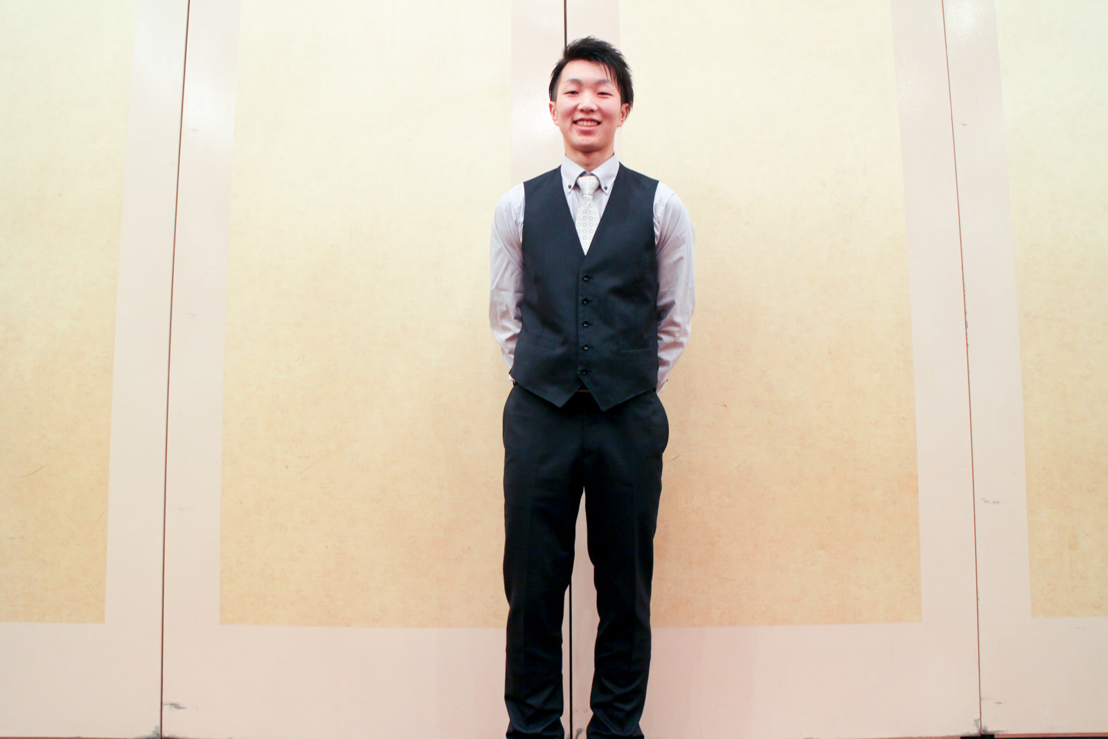 ベージュ色の壁の前で、グレーのシャツに黒のベストとネクタイを身に着けた新成人の男性が両手を後ろで組んで笑顔で立っている写真